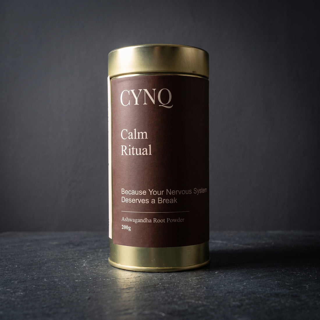 Calm Ritual ashwagandha powder tin, front view, on dark slate against a charcoal wall