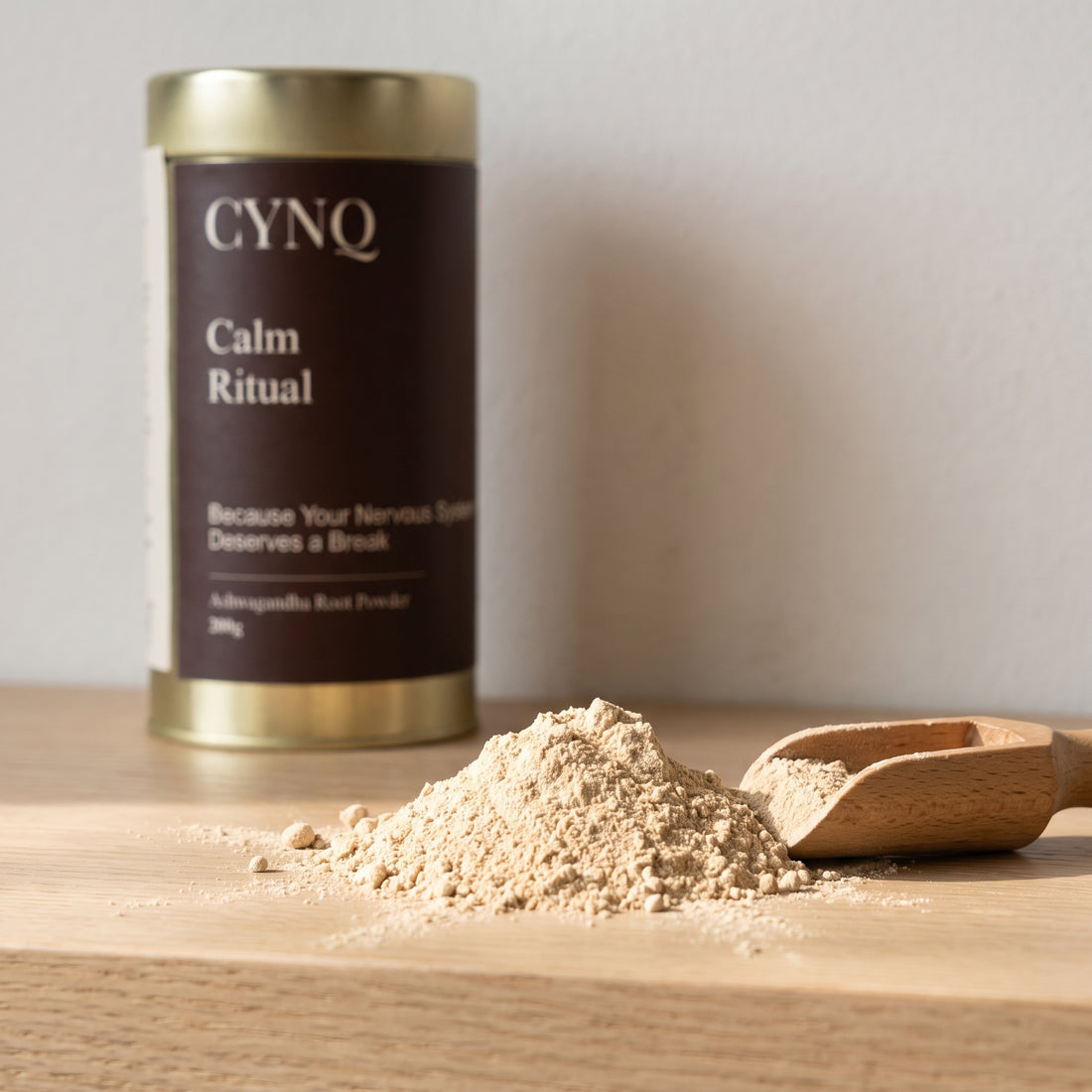Pale ashwagandha root powder with a wooden scoop and a blurred Calm Ritual tin behind