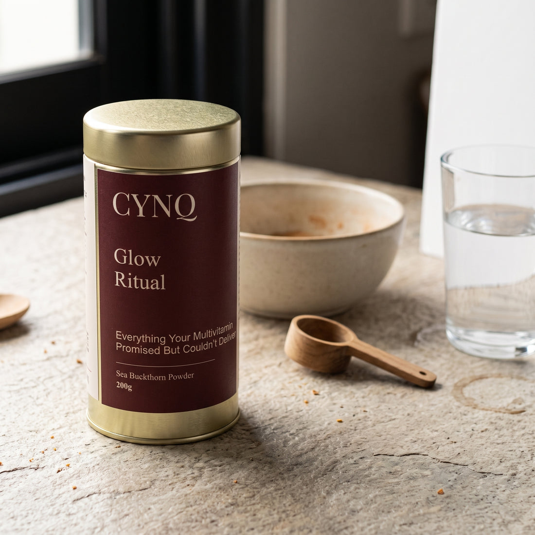 Glow Ritual tin on a stone counter with a stoneware bowl, a wooden scoop and a glass of water