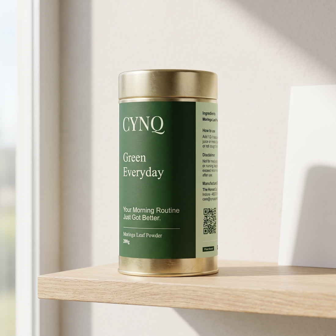 Green Everyday moringa leaf powder in a gold tin with a dark green CYNQ label, on an oak shelf by a window
