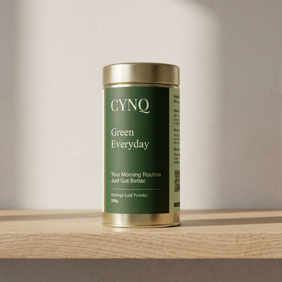 Green Everyday moringa powder tin, front view, on an oak shelf against a pale wall