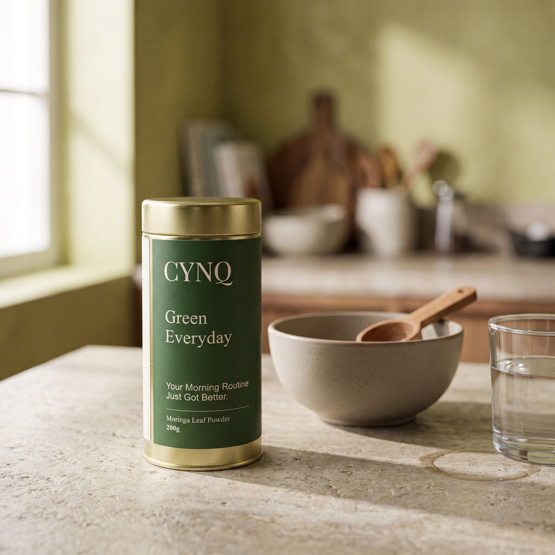 Green Everyday tin on a kitchen counter with a stoneware bowl, a wooden spoon and a glass of water