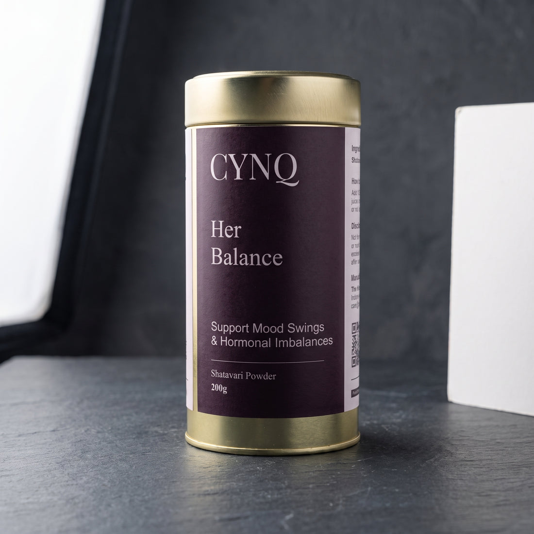 Her Balance shatavari root powder in a gold tin with a plum CYNQ label, on dark slate beside a white card