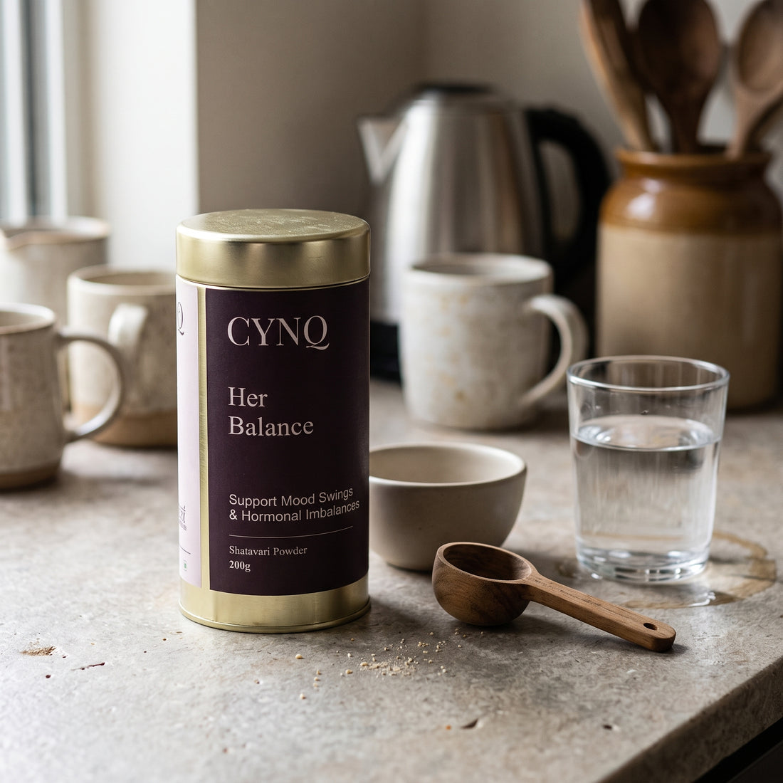 Her Balance tin on a kitchen counter with mugs, a kettle, a wooden spoon and a glass of water