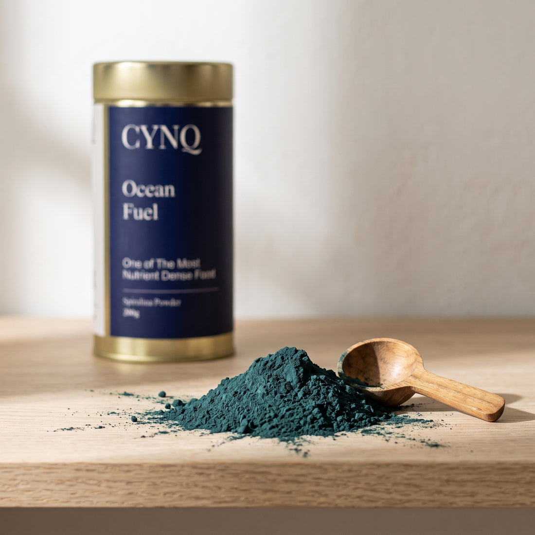 Dark green spirulina powder heaped beside a wooden scoop, with a blurred Ocean Fuel tin behind