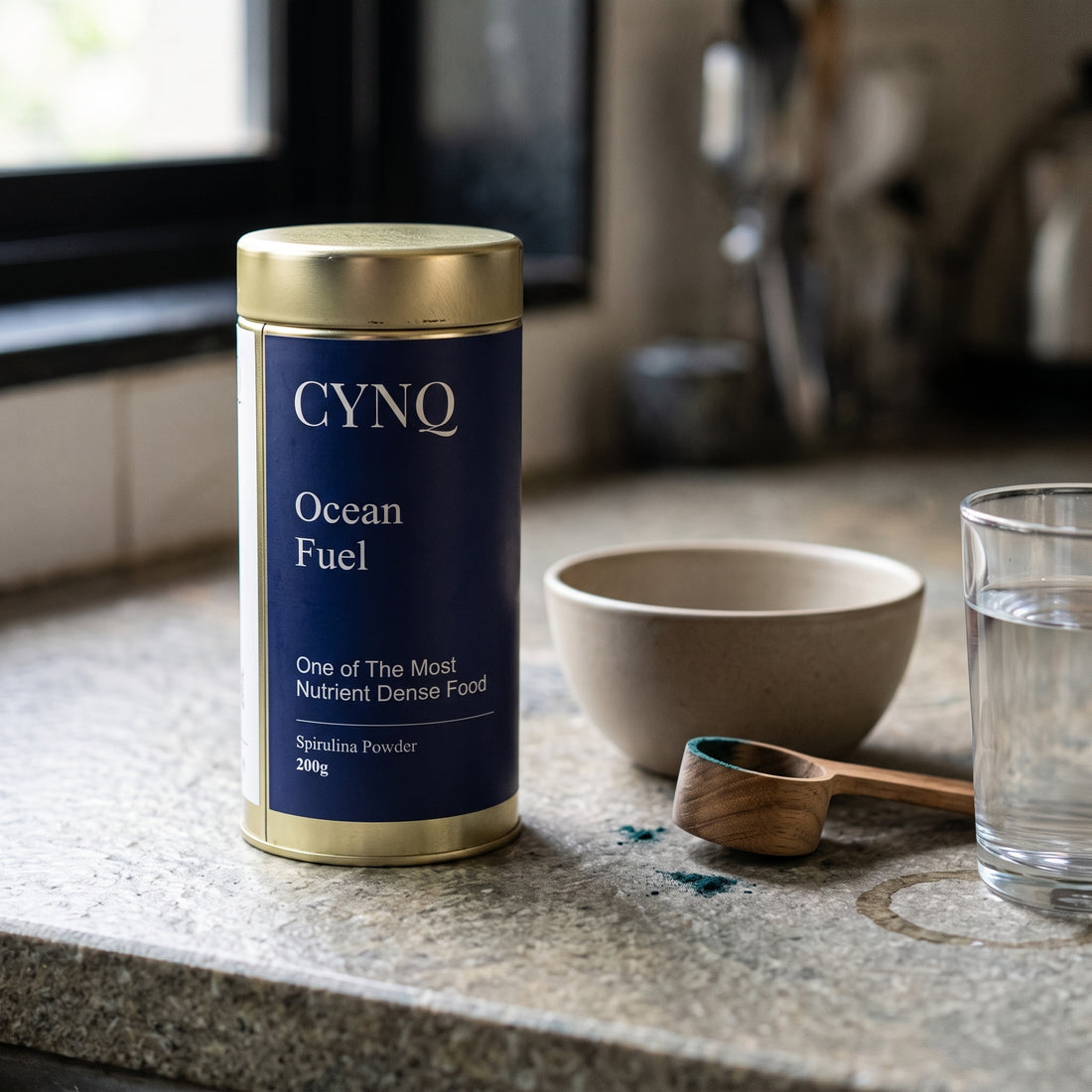 Ocean Fuel tin on a granite counter with a stoneware bowl, a wooden scoop and a glass of water