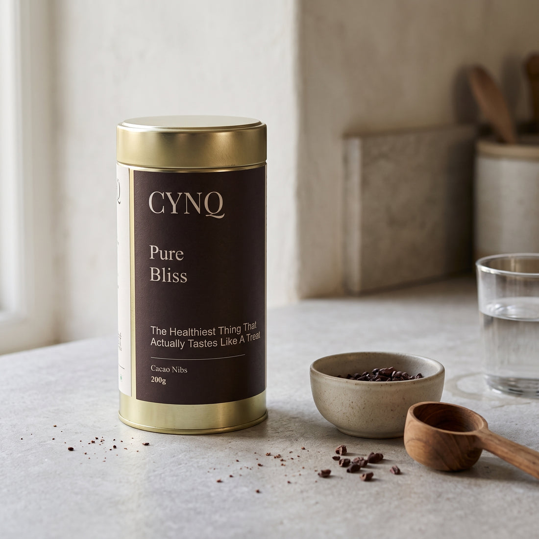Pure Bliss tin on a kitchen counter with a bowl of cacao nibs, a wooden scoop and a glass of water